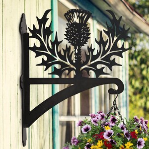 May include: Black metal hanging planter bracket with a thistle design. The bracket is attached to a wooden wall and has a chain hanging from it. A flower pot with colorful flowers is hanging from the chain.