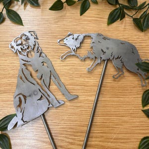 May include: Two metal dog silhouettes on stakes. One dog is sitting, the other is running. The dogs are made of silver metal and are designed to be placed in a garden or yard. Green leaves are in the background.
