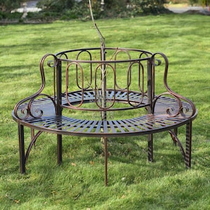 May include: A circular, bronze metal tree seat with a slatted design. The seat has a decorative, curved back and surrounds a young tree. The bench is set on a grassy lawn. The diameter is approximately 120 cm.