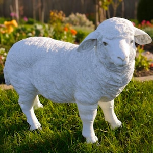 May include: A white sheep statue with a textured wool-like finish stands on green grass. The sheep has a detailed face and ears, and is positioned in a garden setting with flowers in the background. A decorative garden ornament.