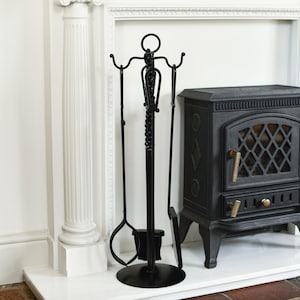May include: Black fireplace tool set with a stand, including a poker, shovel, and tongs. The tools have decorative elements and are displayed near a black cast iron stove. The set is made of metal and is designed for use with a fireplace.