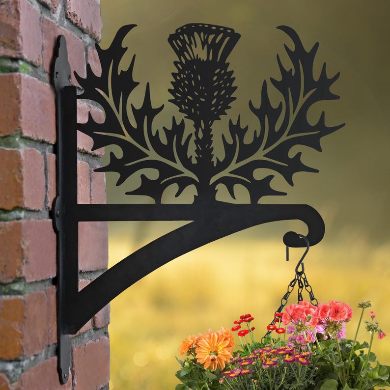 May include: Black metal plant hanger with a thistle design. The hanger is attached to a brick wall and has a hanging basket with colorful flowers.