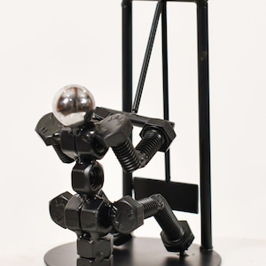 May include: A metal sculpture of a figure performing a lat pulldown exercise, constructed from black nuts, bolts, and screws. The figure has a polished silver ball for a head and is positioned on a circular base in front of a black metal frame.