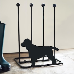 May include: Black metal boot rack with a dog silhouette. The rack has four vertical bars topped with round finials and a square base. A black silhouette of a dog is in the center. A teal wellington boot is on the left.