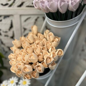 May include: A bouquet of cream-coloured wooden roses in a white vase. The roses are arranged in a circular pattern and are all facing the same direction. The vase is sitting on a white shelf.