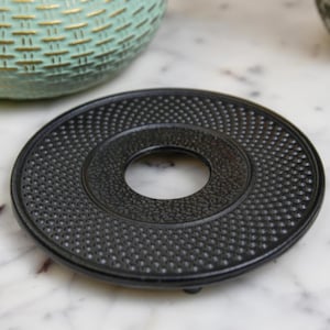 May include: Black cast iron tea cosy with a textured surface and a round hole in the centre. The cosy has three small feet on the bottom.