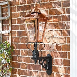 May include: A copper outdoor wall lantern with a glass enclosure and a black metal bracket. The lantern is mounted on a red brick wall. The lantern's design features a pointed top and decorative accents.