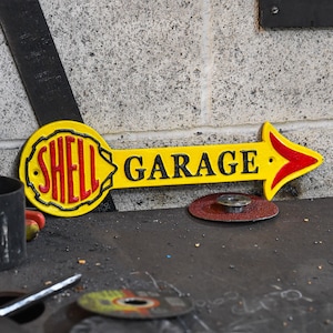 May include: Yellow and red cast iron sign with an arrow pointing to the right. The sign says "Shell Garage".