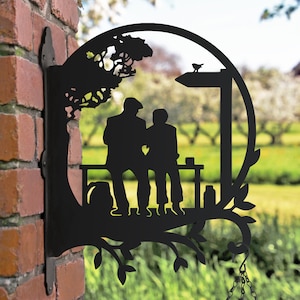 May include: Black metal silhouette of two people sitting on a bench under a tree. The silhouette is attached to a wall with a chain hanging from the bottom.