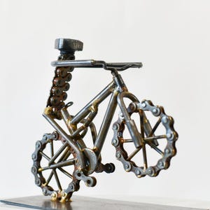 May include: A metal sculpture of a bicycle constructed from recycled industrial parts including steel chains, nuts, and bolts. The frame is made of welded metal rods, with circular chain links forming the wheels and a single nut serving as the seat.