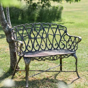 May include: Bronze and black metal garden bench with a decorative back featuring heart-shaped cutouts. The bench has armrests. It is set on a grassy lawn next to a tree, perfect for outdoor seating.