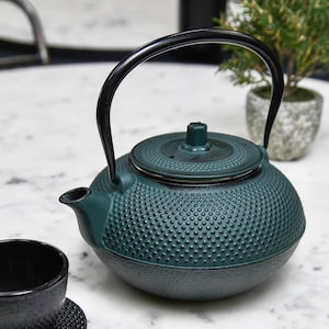 May include: A green cast iron teapot with a black handle and lid. The teapot has a textured surface and is sitting on a white marble surface.