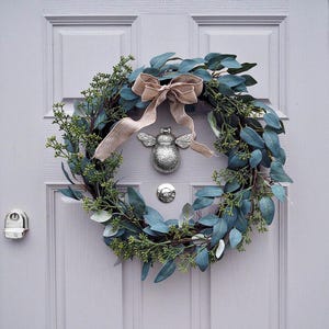 May include: A silver bee door knocker with a brown hessian bow on a green eucalyptus wreath hanging on a light grey door.