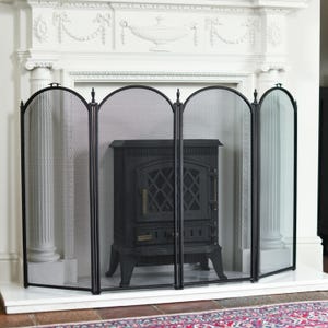 May include: A black metal fireplace screen with a mesh design, protecting a black stove. The screen has four arched panels and stands in front of a white fireplace with decorative molding and columns. The floor has a patterned rug.