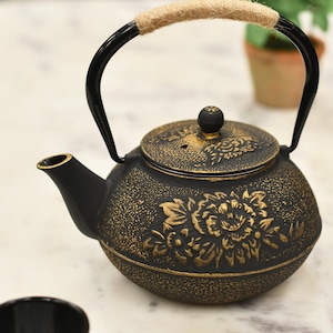 May include: Black cast iron teapot with a gold floral design and a rope handle. The teapot is sitting on a white marble surface.