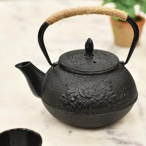 May include: Black cast iron teapot with a floral design and a rope handle. The teapot is sitting on a white marble surface.