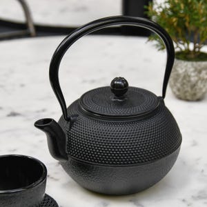 May include: A black cast iron teapot with a textured surface and a curved handle. The teapot has a lid with a small knob and a spout for pouring. The design is a classic style for brewing tea.