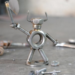 May include: A handcrafted metal sculpture of a warrior figure, constructed from nuts, bolts, and screws. The figure holds a small axe and has screw-shaped horns. The sculpture is silver in color and stands on a metal base.