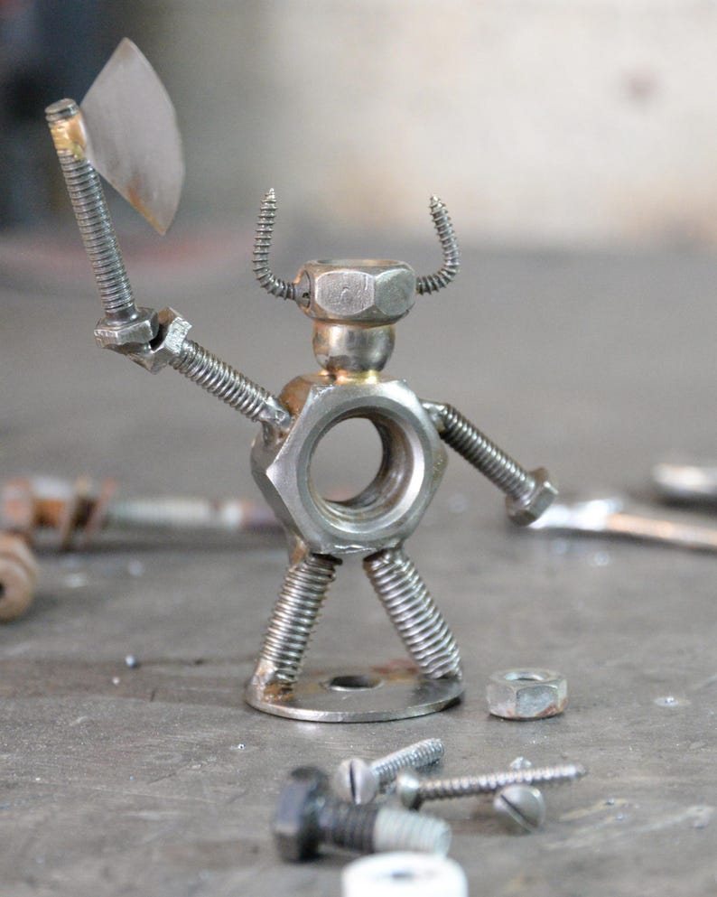 May include: A handcrafted metal sculpture of a warrior figure, constructed from nuts, bolts, and screws. The figure holds a small axe and has screw-shaped horns. The sculpture is silver in color and stands on a metal base.