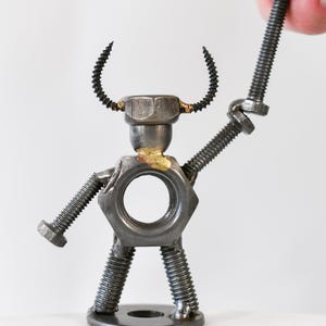 May include: A handcrafted metal sculpture of a figure made from nuts, bolts, and screws. The figure has a hexagonal nut for a torso, a bolt for each arm, and screw legs. A hand holds a bolt above the figure's head.