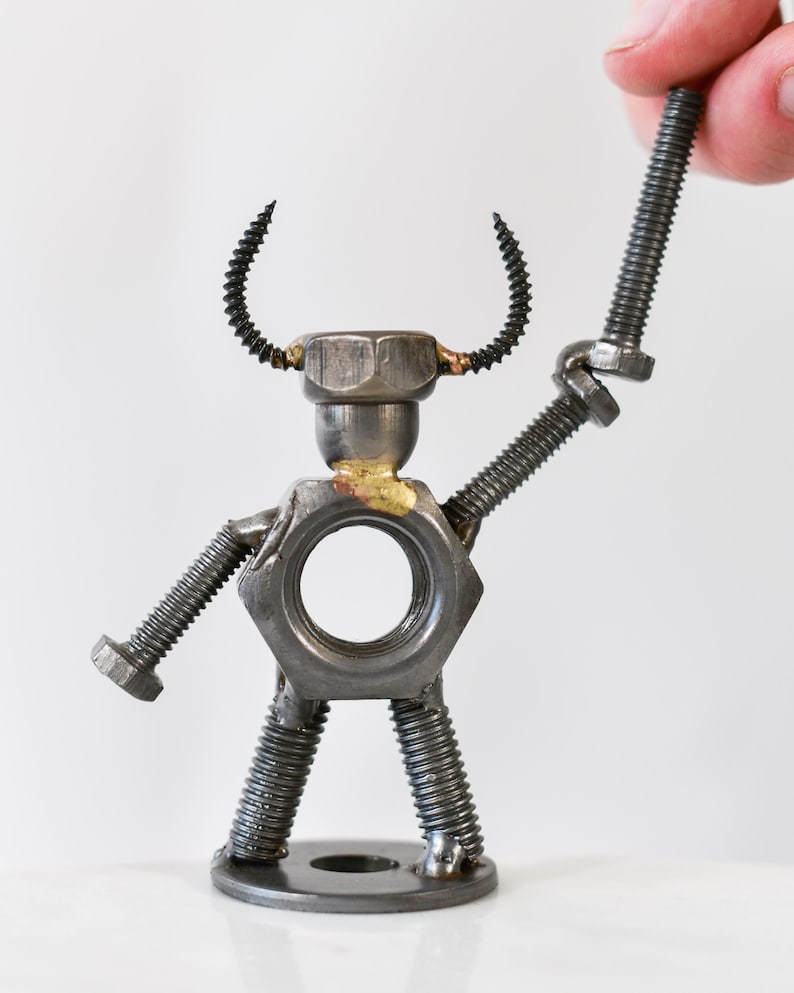 May include: A handcrafted metal sculpture of a figure made from nuts, bolts, and screws. The figure has a hexagonal nut for a torso, a bolt for each arm, and screw legs. A hand holds a bolt above the figure's head.