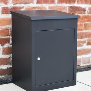 May include: A dark grey metal mailbox with a hinged door and a keyhole. The rectangular box has a flat top. The mailbox is set against a red brick wall.