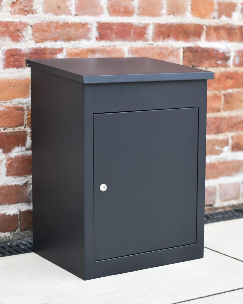 May include: A dark grey metal mailbox with a hinged door and a keyhole. The rectangular box has a flat top. The mailbox is set against a red brick wall.
