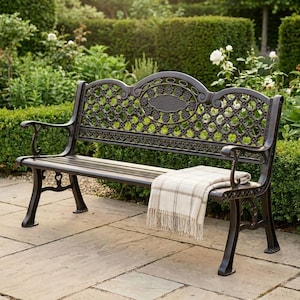 May include: A dark brown metal garden bench with ornate floral and lattice detailing. The bench features armrests and a slatted seat. A beige and white checked blanket is draped over the seat. The bench is set on a stone patio, surrounded by a garden.