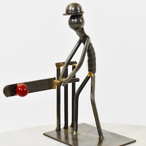 May include: A metal sculpture depicting a cricket player. The figure is crafted from welded metal rods, complete with a helmet, bat, and a red ball. The sculpture is mounted on a rectangular metal base.