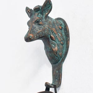 May include: A bronze-effect deer head wall hook with a verdigris finish. The hook is mounted on a white wall and holds a black garment. The deer head has detailed features and a rustic aesthetic.