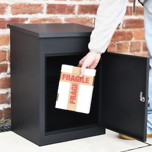 May include: A black parcel box with an open door. A person is placing a white box with "FRAGILE" printed on it inside. The box is made of metal and has a simple, modern design.