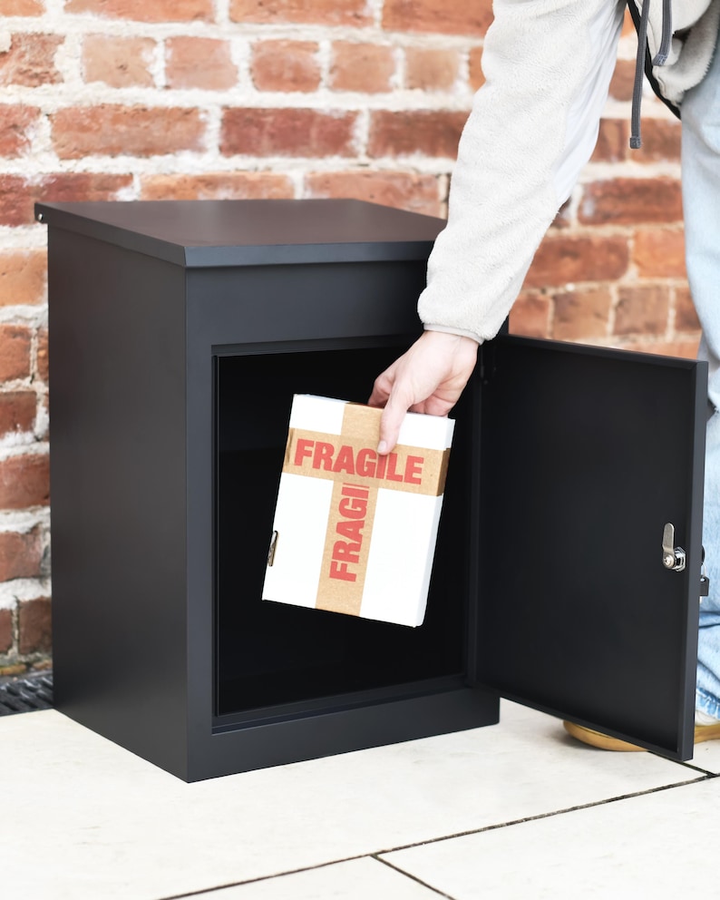 May include: A black parcel box with an open door. A person is placing a white box with "FRAGILE" printed on it inside. The box is made of metal and has a simple, modern design.
