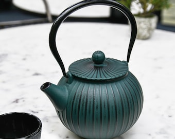 May include: A teal green cast iron teapot with a black handle and lid. The teapot has a ribbed design and is sitting on a white marble surface.