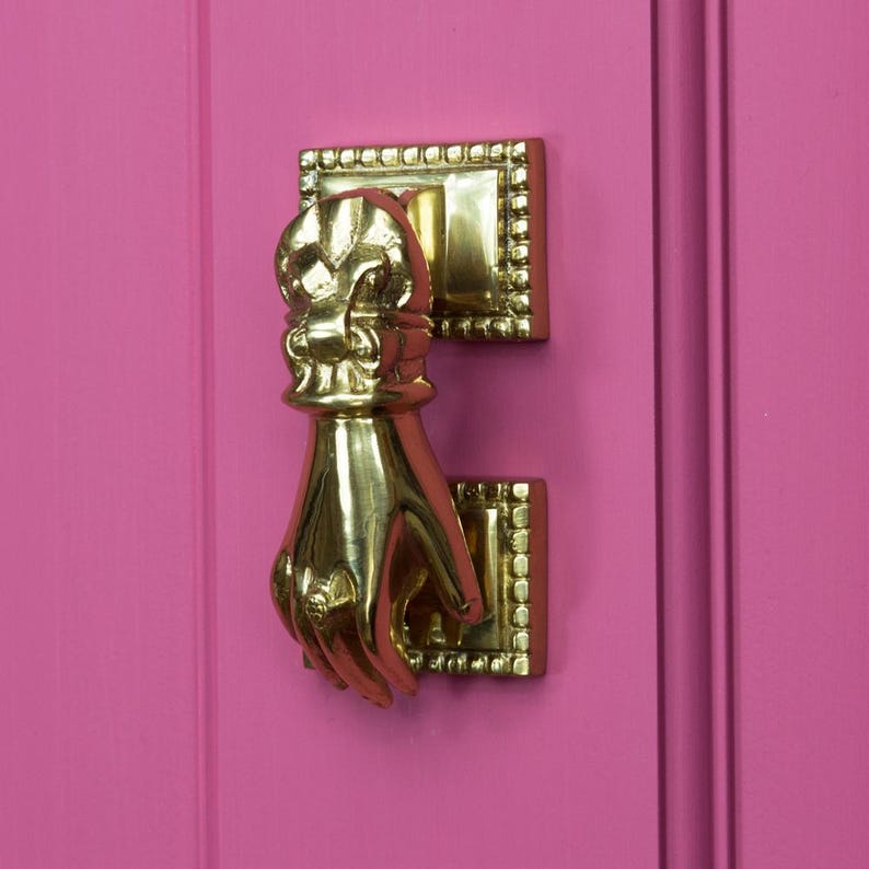 May include: A gold-toned door knocker shaped like a hand with a decorative design. The knocker is mounted on a pink door.