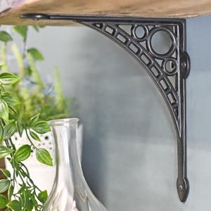May include: A dark grey, decorative shelf bracket with an ornate design. The metal bracket has a curved arm with circles and rectangular cutouts. It is mounted on a wooden shelf and a light blue wall.
