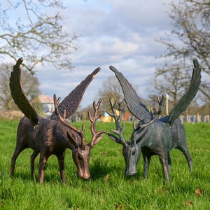 May include: Two bronze deer sculptures with large, detailed wings. The deer have antlers and are positioned on a grassy lawn under a cloudy sky. The sculptures are detailed and artistic.