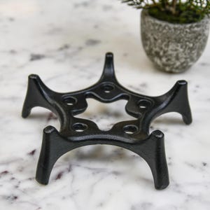 May include: A black cast iron trivet with a starburst design. The trivet has six curved legs and a series of circular openings. The trivet is designed to protect surfaces from hot cookware. A small potted plant is in the background.