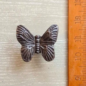 May include: A silver metal butterfly-shaped drawer pull. The pull is approximately 2.5 cm wide.