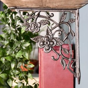 May include: A dark metal shelf bracket with a floral design, including flowers and leaves. The bracket supports a wooden shelf. The background includes greenery and a red book, adding a touch of colour.