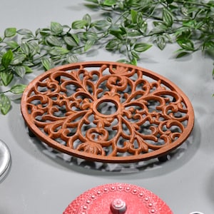 May include: Oval, rust-coloured cast iron trivet with an intricate, openwork floral design. The trivet has a central circular opening and small feet. Decorative kitchenware.