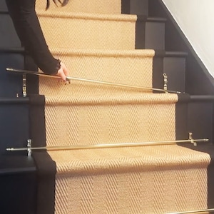 May include: A set of gold metal stair rods with decorative finials, installed on a set of stairs with black painted risers and natural fibre carpet.