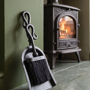 May include: A black metal fireplace dustpan and brush set featuring a hand-forged handle with a decorative scrolled loop design. The dustpan has a wide, flat base, and the brush is equipped with dense, stiff black bristles for cleaning ash.