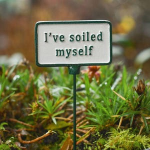 May include: A rectangular garden sign with a white background and a dark green border, mounted on a thin green metal stake. The sign features the embossed dark green text "I've soiled myself" in a clear, sans-serif font, centred on the face.