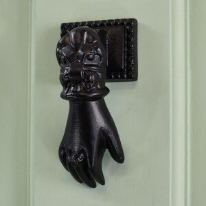 May include: Black cast iron door knocker shaped like a hand with a decorative floral design on the back of the hand.