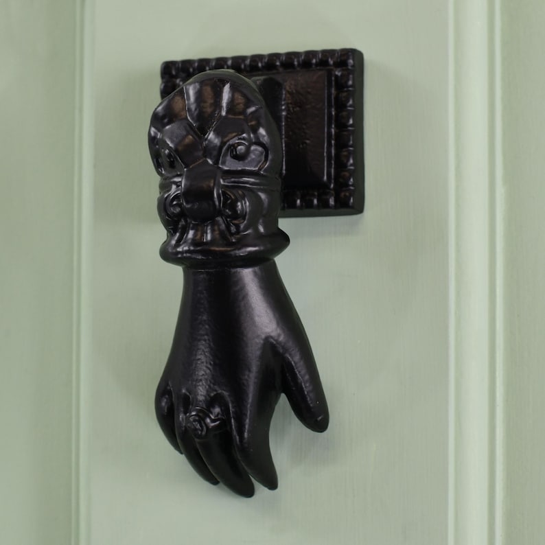 May include: Black cast iron door knocker shaped like a hand with a decorative floral design on the back of the hand.