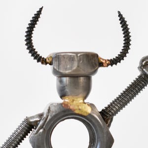 May include: A metal sculpture of a figure made from nuts, bolts, and screws. The head is a hex nut with curved screw horns. The arms are made of screws, and the body has a large hole.