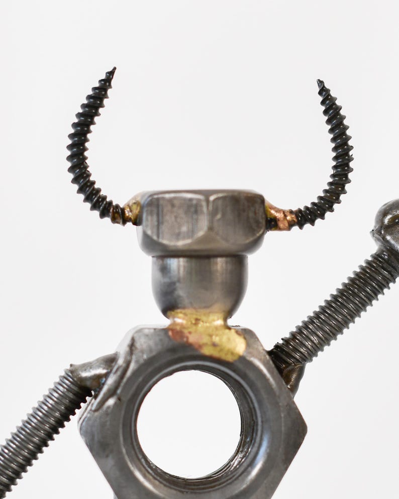 May include: A metal sculpture of a figure made from nuts, bolts, and screws. The head is a hex nut with curved screw horns. The arms are made of screws, and the body has a large hole.
