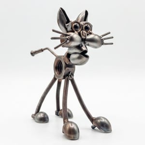 May include: A cat figurine constructed from repurposed metal hardware. The body features a large hexagonal nut, while the legs are made of coiled springs ending in rounded metal feet. The head includes dark circular eyes and metal whiskers.