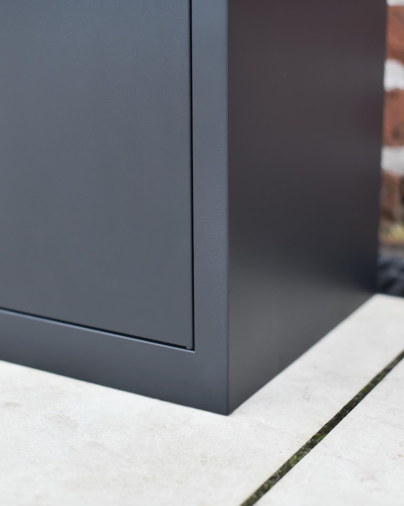 May include: A close-up of a dark grey, rectangular cabinet. The cabinet has a simple, modern design with clean lines and a matte finish. The cabinet is sitting on a light-coloured, textured surface.