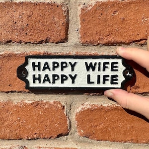 May include: A rectangular black and white metal sign with rounded ends and two mounting holes. The sign features raised black lettering on a textured white background that reads, "HAPPY WIFE HAPPY LIFE" in two lines of bold, capitalized text.
